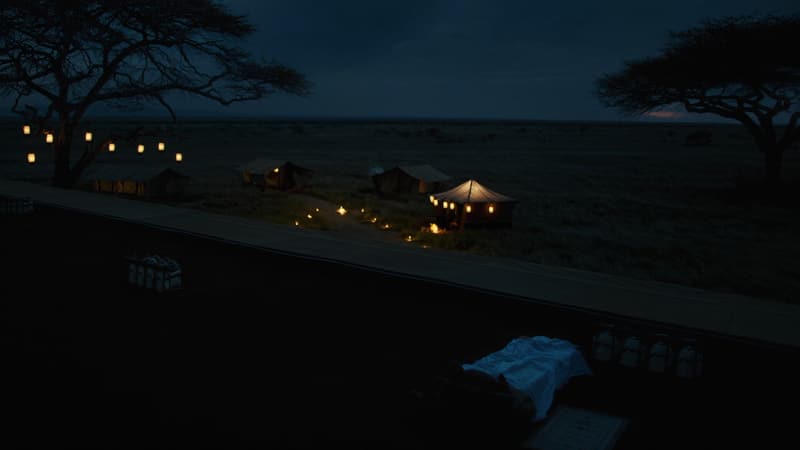 Kambi ya safari yenye taa za manjano ikiwaka jioni kwenye uwanda wa Serengeti